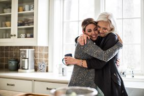 A mother and adult daughter hug in kitchen
