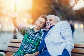 Preteen boy takes selfie with older man on park bench.